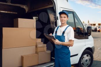 Deliveryman in uniform, carton boxes in the car, delivery service. Man standing at cardboard packages in vehicle, male deliver, courier or shipping job