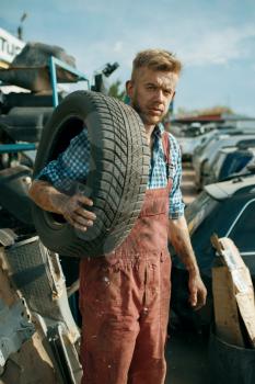 Male repairman holds tire on car junkyard. Auto scrap, vehicle junk, automobile garbage, abandoned, damaged and crushed transport
