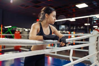 Woman in black boxing bandages and sportswear poses at the ropes on the ring, box training. Female boxer in gym, girl kickboxer in sport club, punches practice