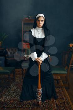 Young nun in a cassock holds a baseball bat wrapped in a chain, vicious desires. Corrupt sister in the monastery, religion and faith, sinful religious people, attractive sinner