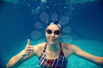 Female swimmer in swimsuit, swimming cap and glasses shows thumbs up underwater in pool. The woman holds her breath in the water, breath-hold technique exercise