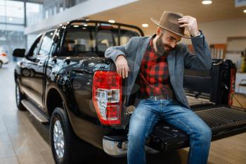 Happy man poses in the back of new pickup truck in car dealership. Customer in vehicle showroom, male person buying transport, auto dealer business