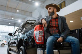 Man in hat poses at new pickup truck in car dealership. Customer in vehicle showroom, male person buying transport, auto dealer business