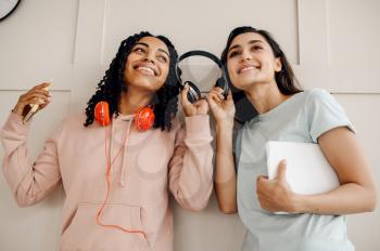 Two smiling women in headphones listening to music indoors. Pretty girlfriends in earphones relax in the room, music lovers resting