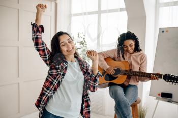 Funny women play the guitar and dancing at home. Pretty girlfriends in earphones relax in the room, sound lovers resting on couch, female friends leisures together
