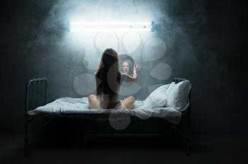 Mad woman with mirror sitting in bed, insomnia, dark room on background. Psychedelic female person having problems every night, depression and stress, sadness, psychiatry hospital