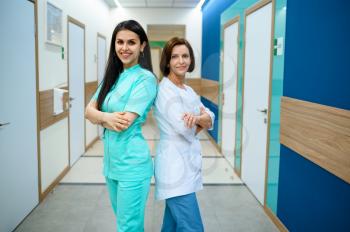 Two smiling female doctors in uniform poses in clinic corridor. Professional medical specialist in hospital, laryngologist or otolaryngologist, gynecologist or mammologist, surgeon