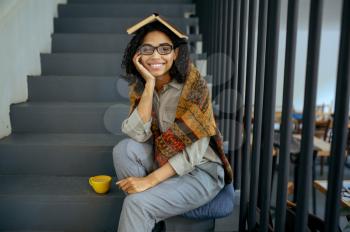 Pretty female student with book sitting on the steps in library cafe. Woman learning a subject, education and knowledge. Girl studying in campus