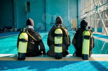 Instructor and two divers in suits sitting at the poolside, back view, diving school. Teaching people to swim underwater with scuba gear, indoor swimming pool interior on background, group training