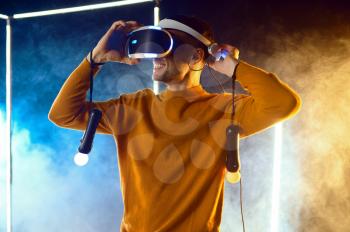Young man plays the game using virtual reality headset and gamepad in luminous cube. Dark playing club interior, spotlight on background, VR technology with 3D vision