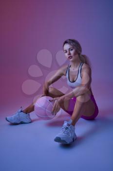 Young sexy sportswoman with ball sitting on the floor in studio, neon background. Fitness woman at the photo shoot, sport concept, active lifestyle motivation