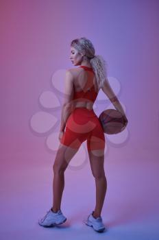 Sexy female athlete in red sportswear poses with ball in studio, back view, neon background. Fitness sportswoman at the photo shoot, sport concept, active lifestyle motivation