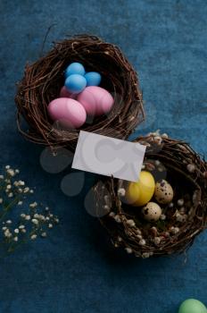 Easter eggs in decorative nests and greeting card on blue cloth background. Paschal food, event decoration, spring holiday celebration symbol