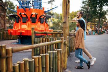 Love couple resting in amusement park, attraction on background. Man and woman relax outdoors. Family leisures in summertime, entertainment theme