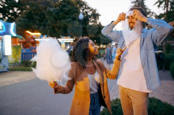 Love couple jokes with cotton candy in city amusement park. Man and woman relax outdoors. Family leisures in summertime, entertainment theme