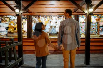 Love couple in amusement park souvenir shop. Man and woman relax outdoors, fairground on background. Family leisures on carousels, entertainment theme