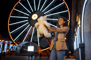 Happy woman hugs soft toy, romantic walking in night amusement park. Family relax outdoors, ferris wheel with lights on background. Love couple leisures on carousels, entertainment theme