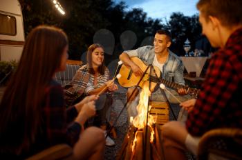 Friends playing on guitar by the bonfire, picnic at camping in the forest. Youth having summer adventure on rv, camping-car on background. Two couples leisures, travelling with trailer