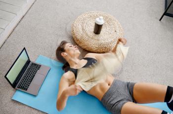 Girl leisures at the laptop after online fit training. Female person in sportswear, internet sport workout, room interior on background