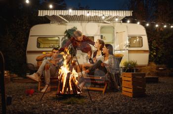 Friends cooking on campfire, picnic at camping in the forest. Youth having summer adventure on rv, camping-car on background. Two couples leisures, travelling with trailer