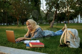 Arab female student in hijab lying on the grass with laptop in summer park. Muslim woman with books resting on the lawn. Religion and education