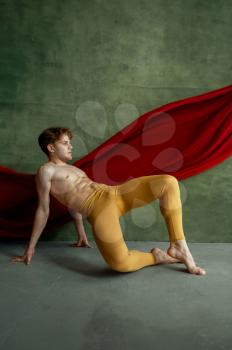 Male ballet dancer doing exercise in dancing studio, grunge wall and red cloth on background. Performer with muscular body, grace and elegance of movements