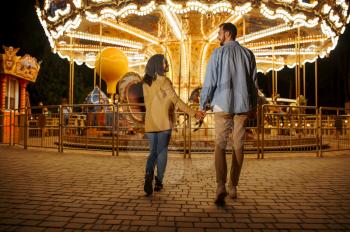Love couple at the carousel in night amusement park. Man and woman relax outdoors, roundabout attraction with lights on background. Family leisures in summertime, entertainment theme