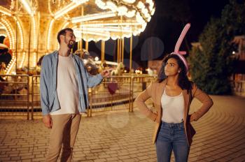 Funny love couple joking in night amusement park. Man and woman relax outdoors, roundabout attraction with lights on background. Family leisures in summertime, entertainment theme