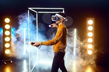 Male gamer plays the game using virtual reality headset and gamepad in luminous cube. Dark playing club interior, spotlight on background, VR technology with 3D vision