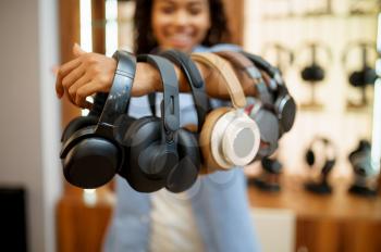 Woman shows headphones collection in audio components store. Female person in music shop, showcase with earphones on background, buyer in multimedia salon