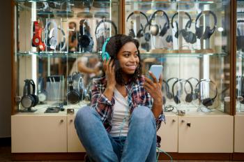 Happy buyer choosing headphones in audio store, music fan. Female person in music shop, showcase with earphones on background, woman in multimedia salon
