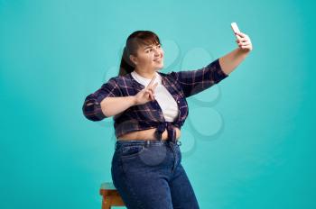 Overweight woman makes selfie in studio, body positive, blue background. Obesity fighting, cheerful female person without complexes