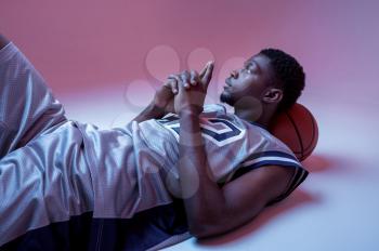 Basketball player lying with ball in studio, neon background. Professional male baller in sportswear playing sport game, tall sportsman