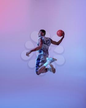 Basketball player jumping with ball in studio, neon background. Professional male baller in sportswear playing sport game, tall sportsman