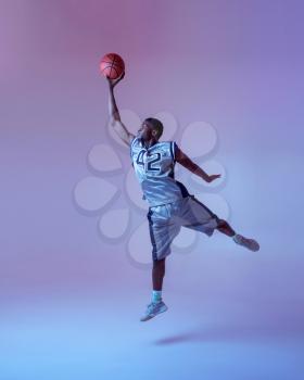 Basketball player with ball shows his skill in studio, jump in action, neon background. Professional male baller in sportswear playing sport game, tall sportsman