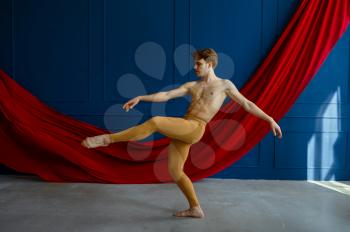Male ballet dancer, training in dancing class, blue walls and red cloth on background. Performer with muscular body, grace and elegance of movements