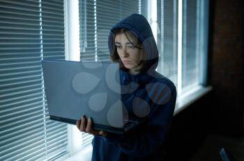 Female internet hacker in hood works on laptop in dark office. Illegal web programmer at workplace, criminal occupation. Data hacking, cyber security