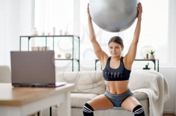 Woman holds ball, online pilates training at the laptop. Female person in sportswear, internet sport workout, room interior on background