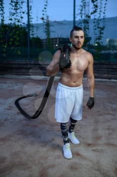 Muscular man poses with crossfit ropes, street workout. Fitness training on sports ground outdoor, male person pumps muscles, active urban lifestyle