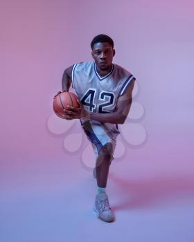 Basketball player practicing with ball in studio, neon background. Professional male baller in sportswear playing sport game.