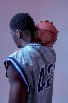 Basketball player poses with ball in studio, back view, neon background. Professional male baller in sportswear playing sport game, tall sportsman