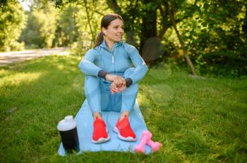 Morning fit training in park, woman in headphones sitting on mat. Female runner goes in for sports at sunny day, healthy lifestyle, jogger on outdoors workout