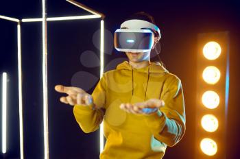 Young gamer plays simulator game in virtual reality headset and gamepad in luminous cube, front view. Dark playing club interior, spotlight on background, VR technology, 3D vision