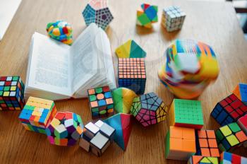 Different puzzle cubes and opened book on the table, nobody. Toy for brain and logical mind training, creative game, solving of complex problems