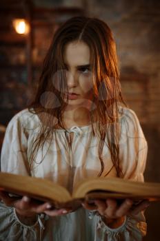 Young demonic woman holds book of spells, demons casting out. Exorcism, mystery paranormal ritual, dark religion, night horror, potions on shelf on background