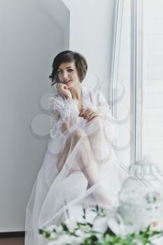 Beautiful young girl in a transparent negligee sitting on a window sill with flowers.