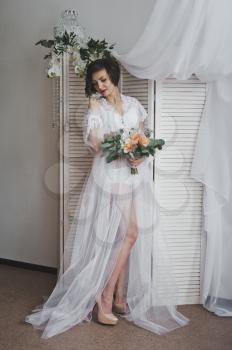 Girl in a semitransparent negligee stands on the background of blinds and curtains.
