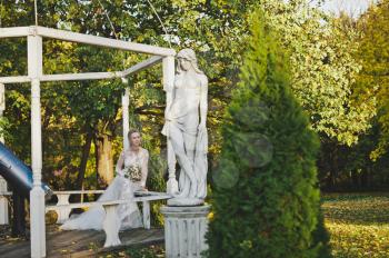 The bride resting on a bench in a beautiful garden with statues.
