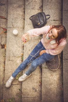 girl is sitting on a skateboard in headphones with a glass of coffee and a smartphone. View from above