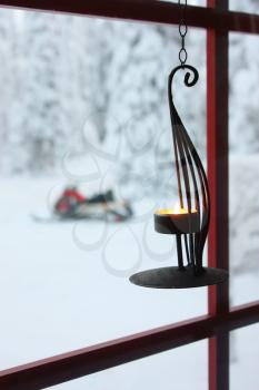 Decorative candle holder with burning candle hanging on window to the snowy winter yard with trees covered in snow and snowmobile